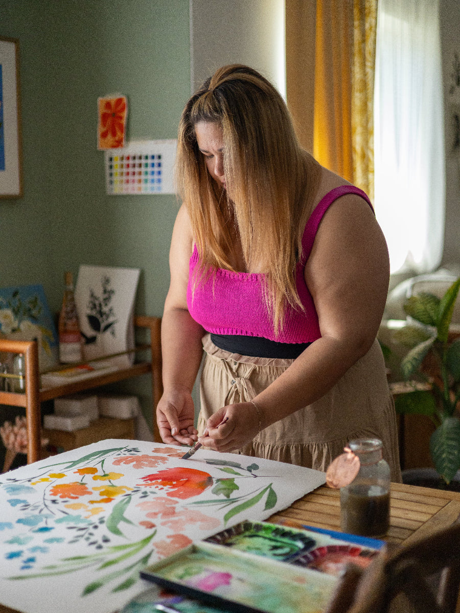 Woman painting a floral scene on paper with a color palette and art supplies around her.
