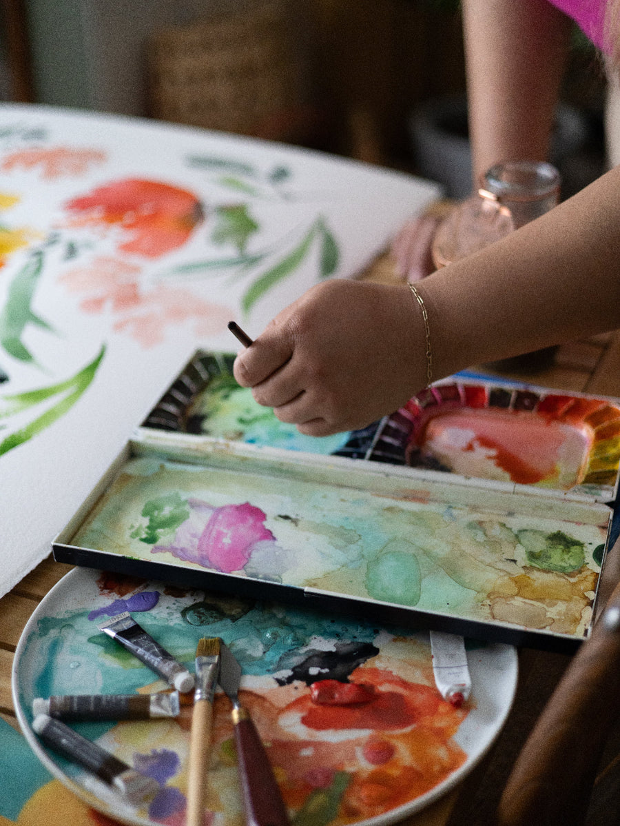 Person painting with watercolors on a table, surrounded by art supplies.