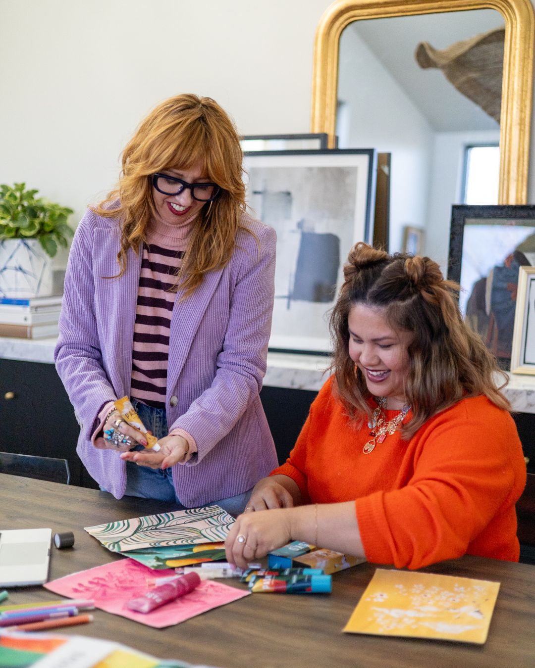 Two women sitting at a table with art supplies, smiling and engaged in a creative activity.
