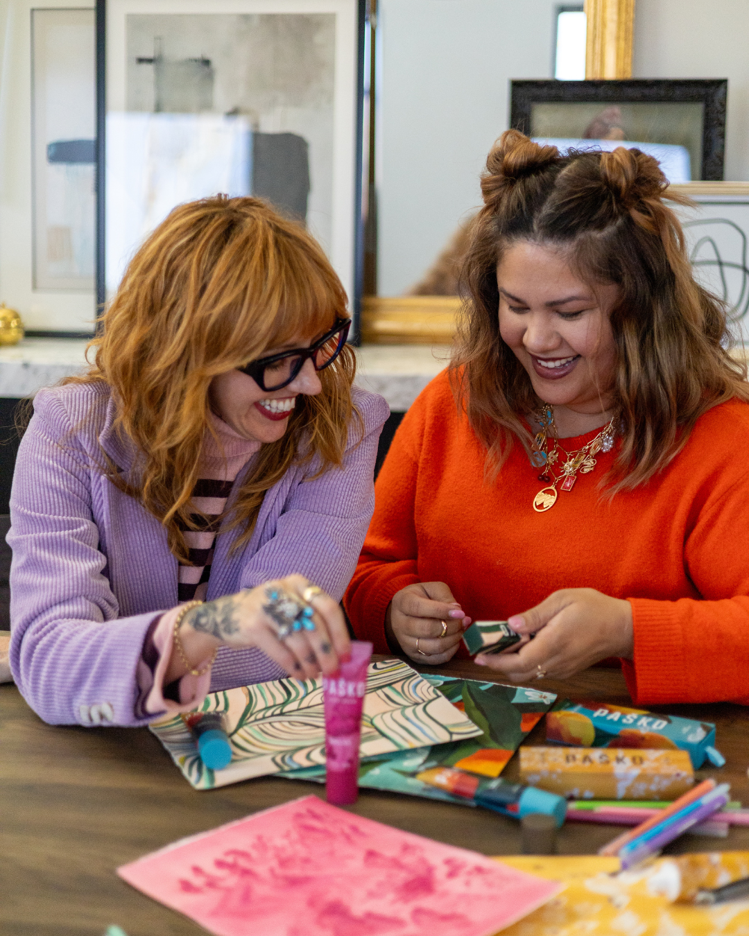 Two women sitting at a table with art supplies, smiling and engaged in a creativing Pasko.