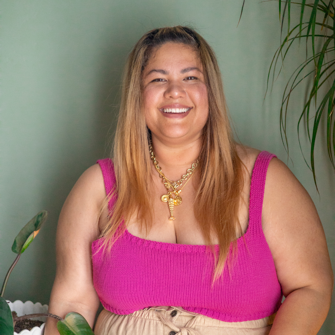 Artist Woman wearing a pink top and beige pants against a green wall with plants.