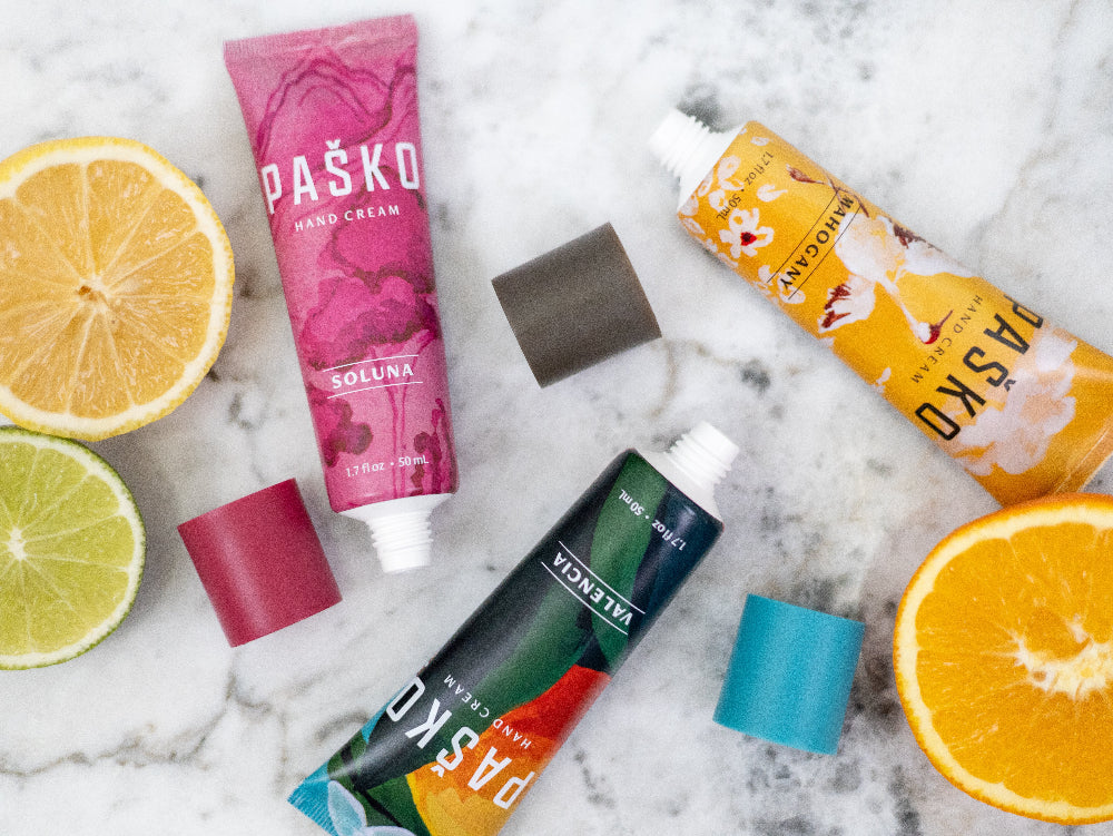 Various tubes of Pasko hand cream with colorful packaging on a marble surface with sliced citrus fruits.